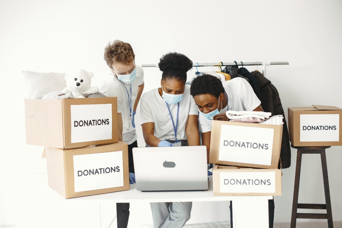 Two smiling girls holding cardboard recycling boxes standing next to a whiteboard with a colored recycling symbol and words glass, plastic, and paper.