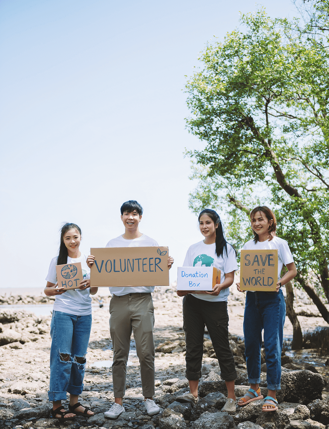 Four young volunteers standing outdoors on rocky ground holding signs that say 'HOT', 'VOLUNTEER', 'Donation Box', and 'SAVE THE WORLD', with a tree and clear sky in the background.