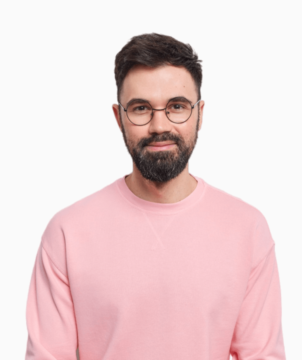 Smiling young man with short curly hair wearing a yellow shirt with rolled-up sleeves and a tattoo on his forearm, standing with arms crossed.