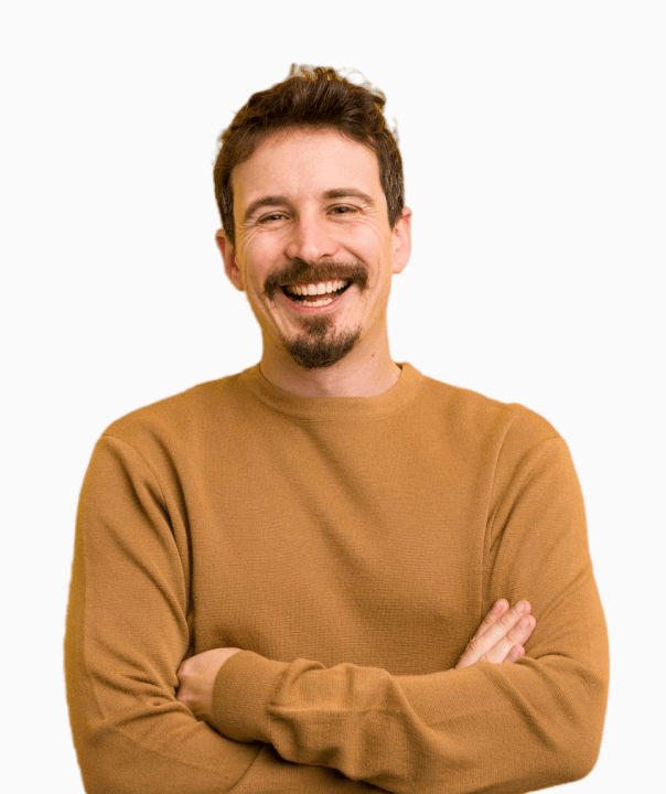 Smiling young man with short curly hair wearing a yellow shirt with rolled-up sleeves and a tattoo on his forearm, standing with arms crossed.