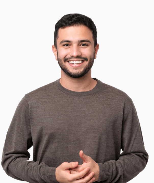 Smiling young man with short curly hair wearing a yellow shirt with rolled-up sleeves and a tattoo on his forearm, standing with arms crossed.