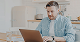 Man in blue shirt working on a laptop at a kitchen table.