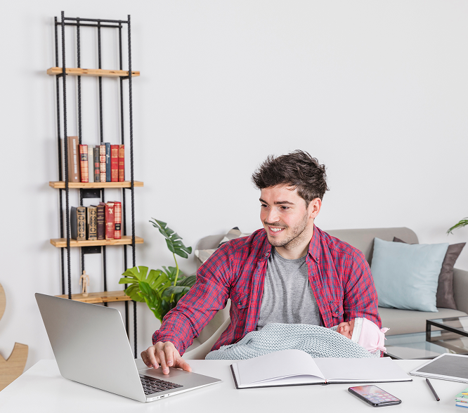 Smiling man holding a baby wrapped in a blanket while using a laptop at a white desk in a cozy living room.