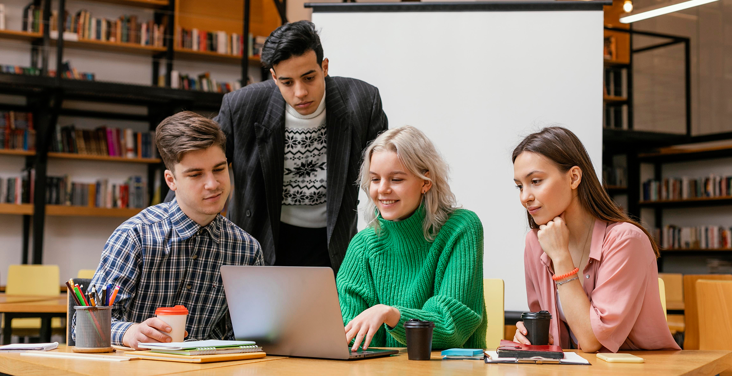 Four young adults collaborating around a laptop in a library with bookshelves in the background.