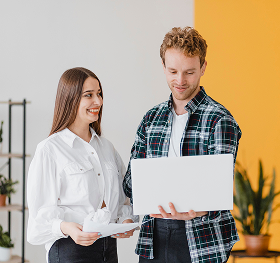 A man and woman standing and looking at a laptop together in a bright room with plants.