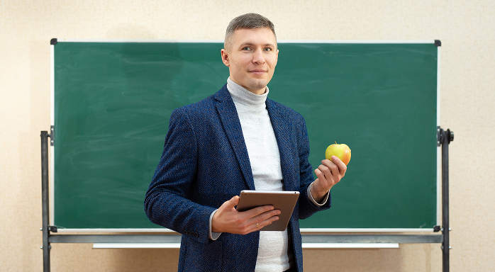 Male teacher in a blue blazer holding a tablet and a green apple standing in front of a blank chalkboard.