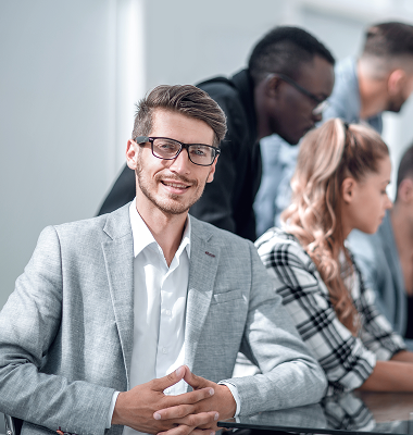 Smiling young man wearing glasses and a light gray suit sitting in front of a group of diverse people in a meeting or classroom setting.