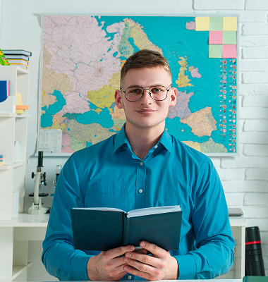 Young man wearing glasses and a blue shirt holding an open book in front of a wall map of Europe.