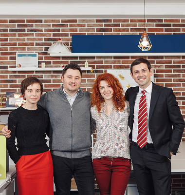 Four colleagues standing closely together and smiling in a modern office with brick wall and shelves.