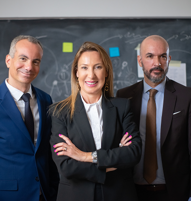 Three confident business professionals standing together in formal attire in front of a chalkboard.