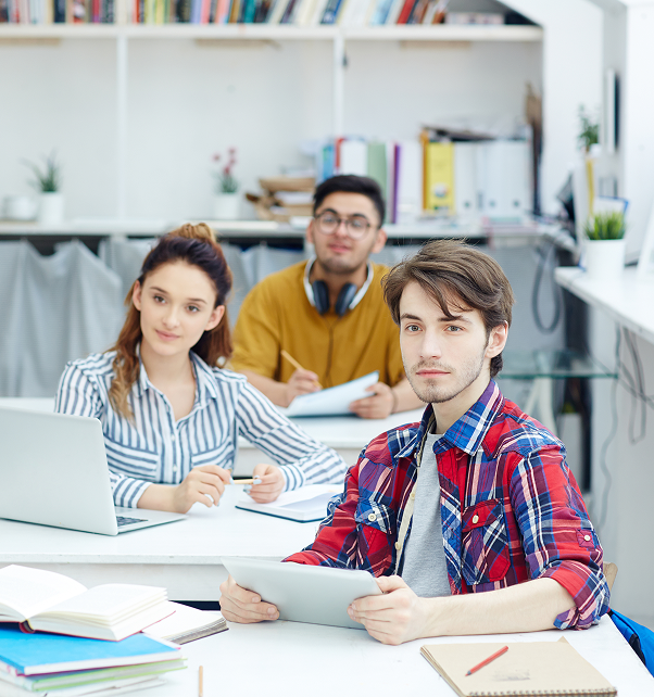 Three young students studying together at a white desk with books, a laptop, and a tablet in a bright room with shelves and plants in the background.