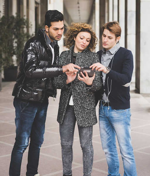 Three young adults standing together outdoors looking at one person's smartphone, engaged in a group activity.