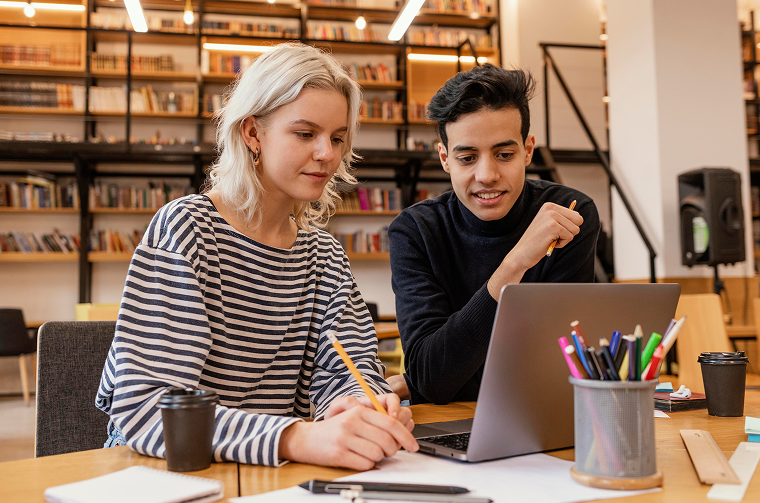 Two young people working together at a table in a library, looking at a laptop and taking notes.